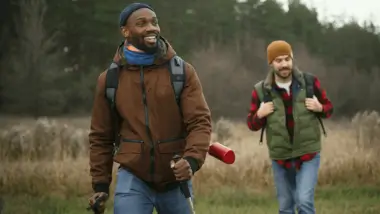 The image features two men walking through a grassy field, each carrying a backpack and a ski pole.